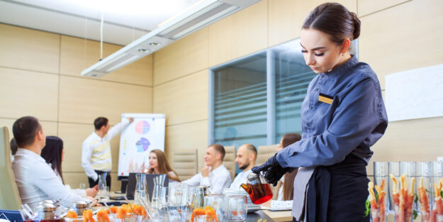 Un'addetta al servizio ristorazione di un Hotel versa dei cocktail, sullo sfondo un tavolo con riunione aziendale